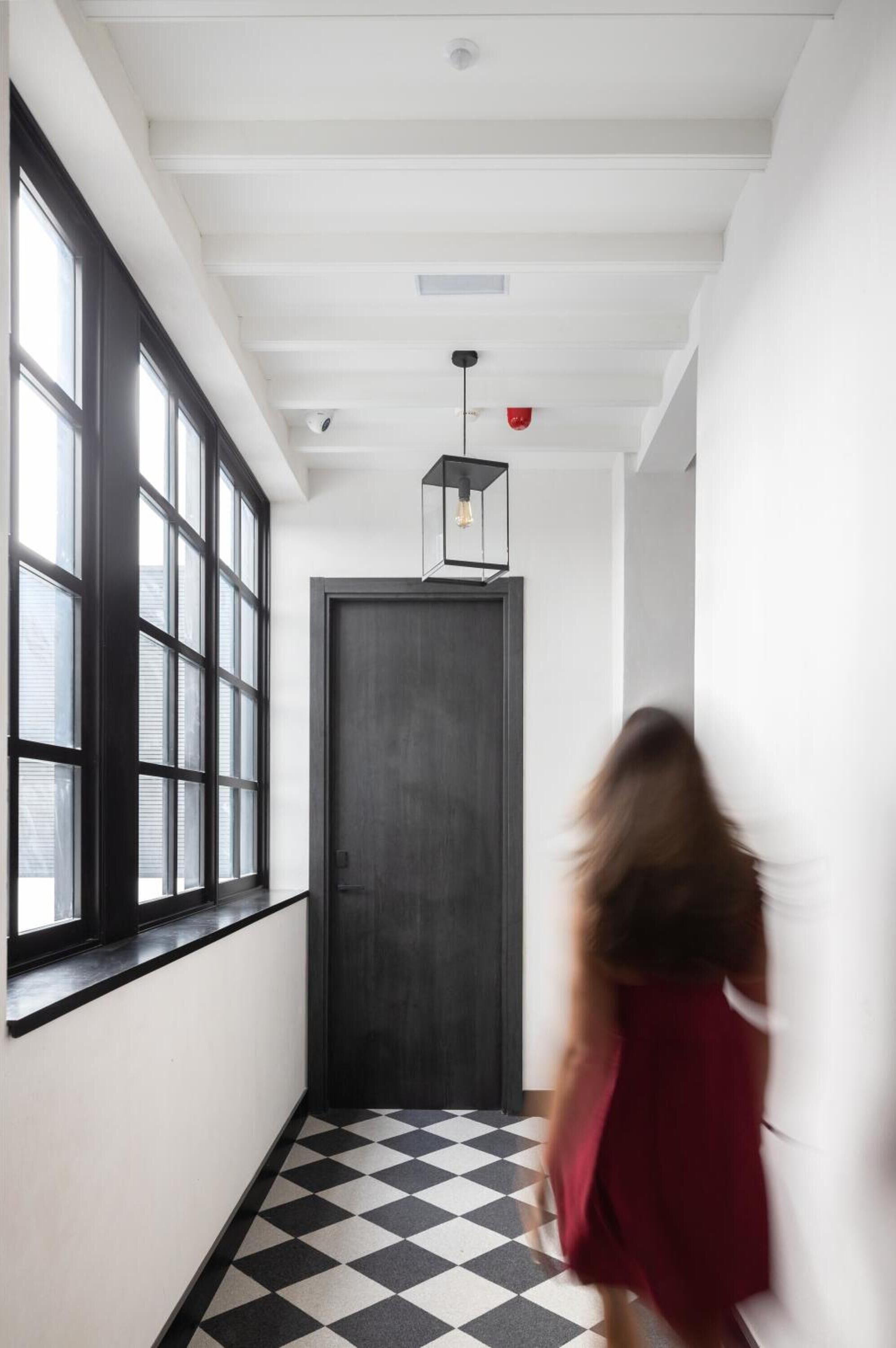 A hallway with large windows, a black and white checkered floor, and a dark door at the end.