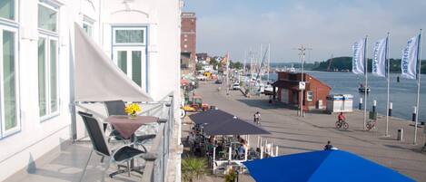 A balcony with outdoor seating, a view of a waterfront street with boats, and a building with a white facade.