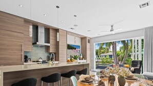 A modern kitchen with a central island, bar stools, and a dining area with a table set for two. - Heated Pool, Modern Spacious Waterway Home Offering Comfort, Style & Total Relaxation! (Wilton Manors)