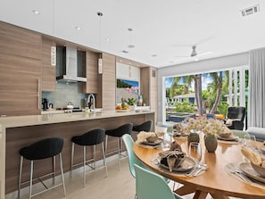 A modern kitchen with a central island, bar stools, and a dining area with a table set for two. - Heated Pool, Modern Spacious Waterway Home Offering Comfort, Style & Total Relaxation! (Wilton Manors)