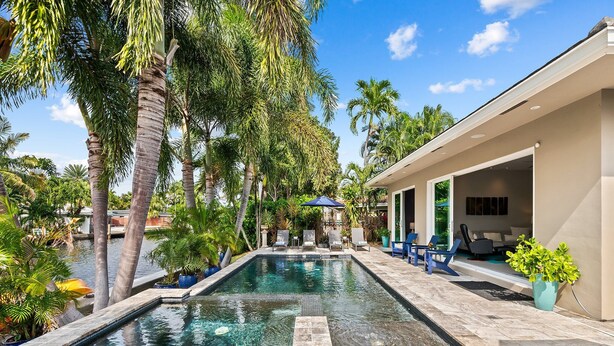 A swimming pool with a stone deck, surrounded by palm trees and a covered seating area with chairs. - Heated Pool, Modern Spacious Waterway Home Offering Comfort, Style & Total Relaxation! (Wilton Manors)