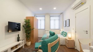 Una habitación compacta y luminosa con cama, televisor, escritorio pequeño y una planta.