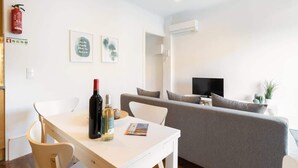 Una sala de estar moderna con un sofá gris, una mesa de comedor blanca con sillas, un televisor y un extintor de incendios en la pared.