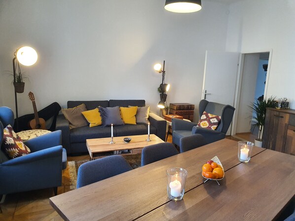 Uma sala de estar com sofá, cadeiras, uma mesa de madeira e uma mesa de jantar. Há velas sobre as mesas.