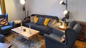 A cozy living room with a dark blue sofa, two armchairs, a wooden coffee table, and a patterned rug. There are two floor lamps and a guitar leaning against the wall. - Central Graz Apartments by Paymán Club (Graz)