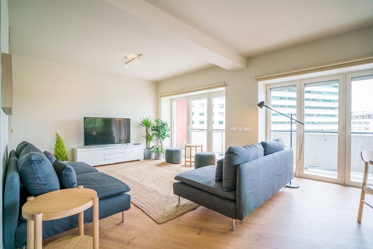 A modern living room with a flat-screen TV, sofas, a coffee table, and a large window with a view of buildings.