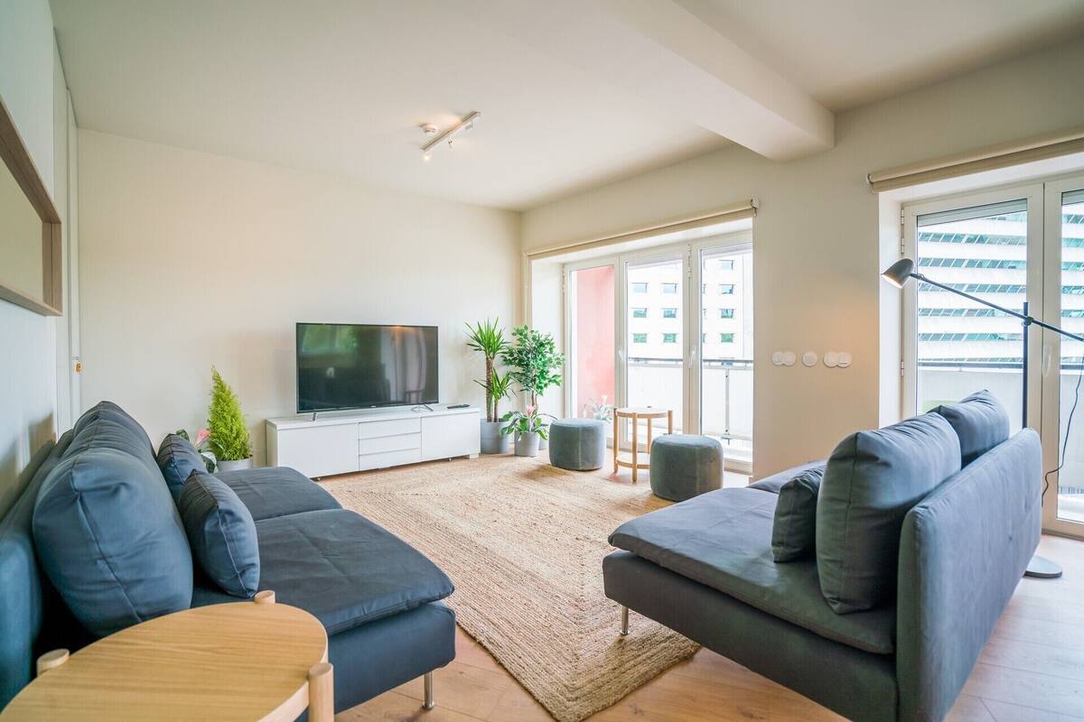 A modern living room with a large flat-screen TV, dark sofas, a wooden coffee table, and a large window with a view of a building.