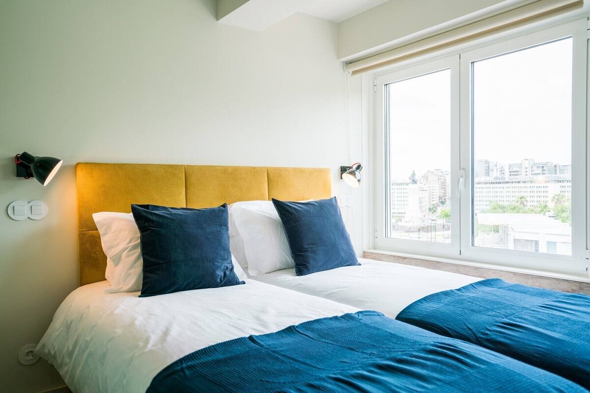 A hotel room with a bed, two bedside lamps, a window with a view of buildings, and a wall-mounted light fixture.