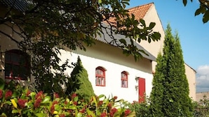 Un bâtiment blanc avec des volets rouges et un toit recouvert de tuiles, entouré de verdure et d’un grand conifère.