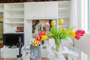 A dining area with a table set for two, a bottle of wine, a vase with tulips, and a television in the background.