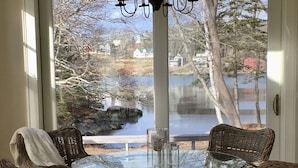 Ein Essbereich mit Korbstühlen, einem Glastisch, einer Chandelier und Blick auf einen See sowie Häuser durch eine Glastür.