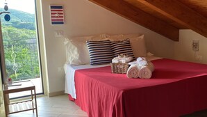 Un dormitorio con una colcha roja, vigas de madera en el techo, un balcón con vistas y una pequeña mesa con una silla.