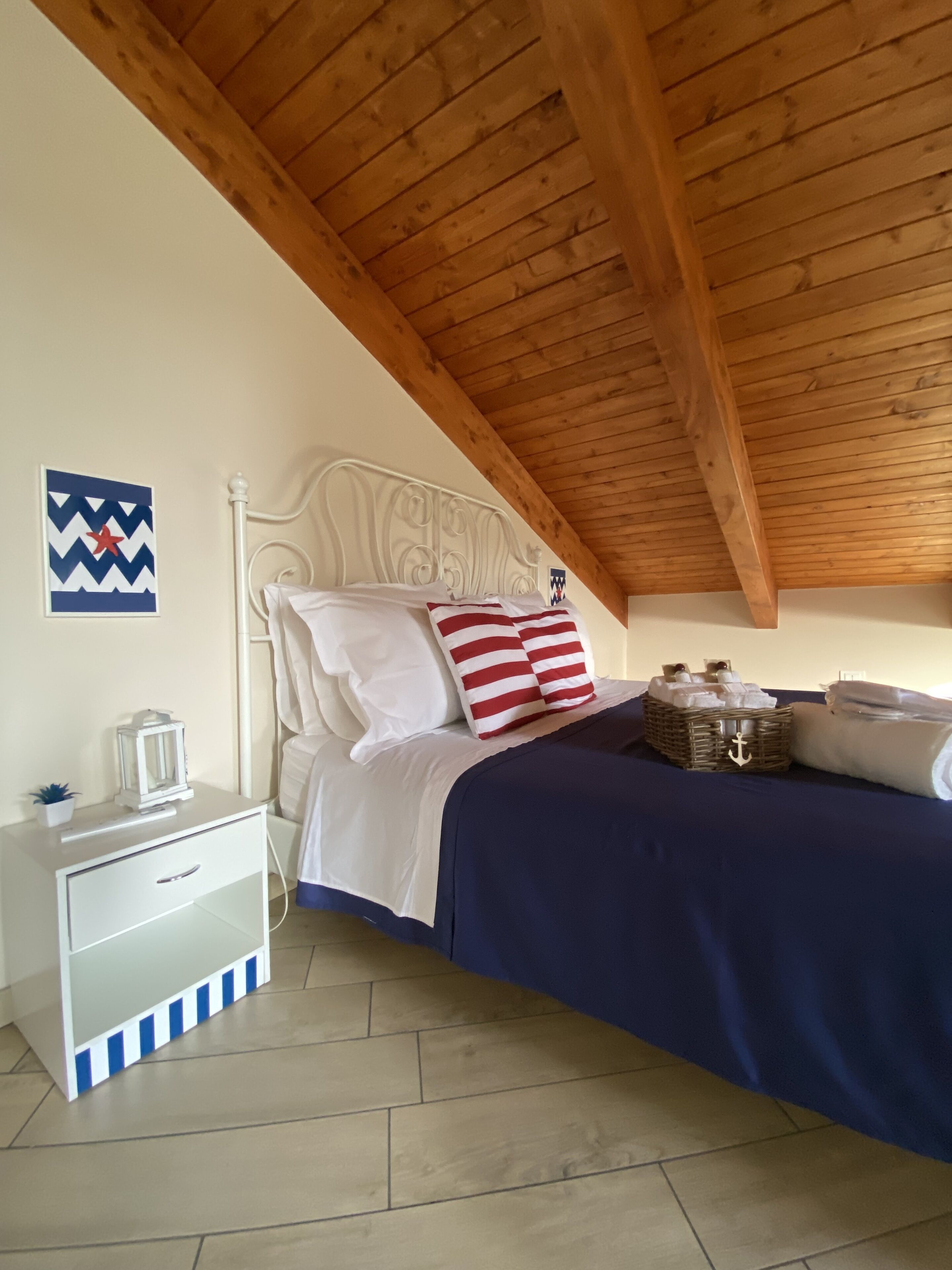 A bedroom with a wooden ceiling, a white bed with navy blue bedding, a white nightstand with a lamp, and a wall with a decorative piece.