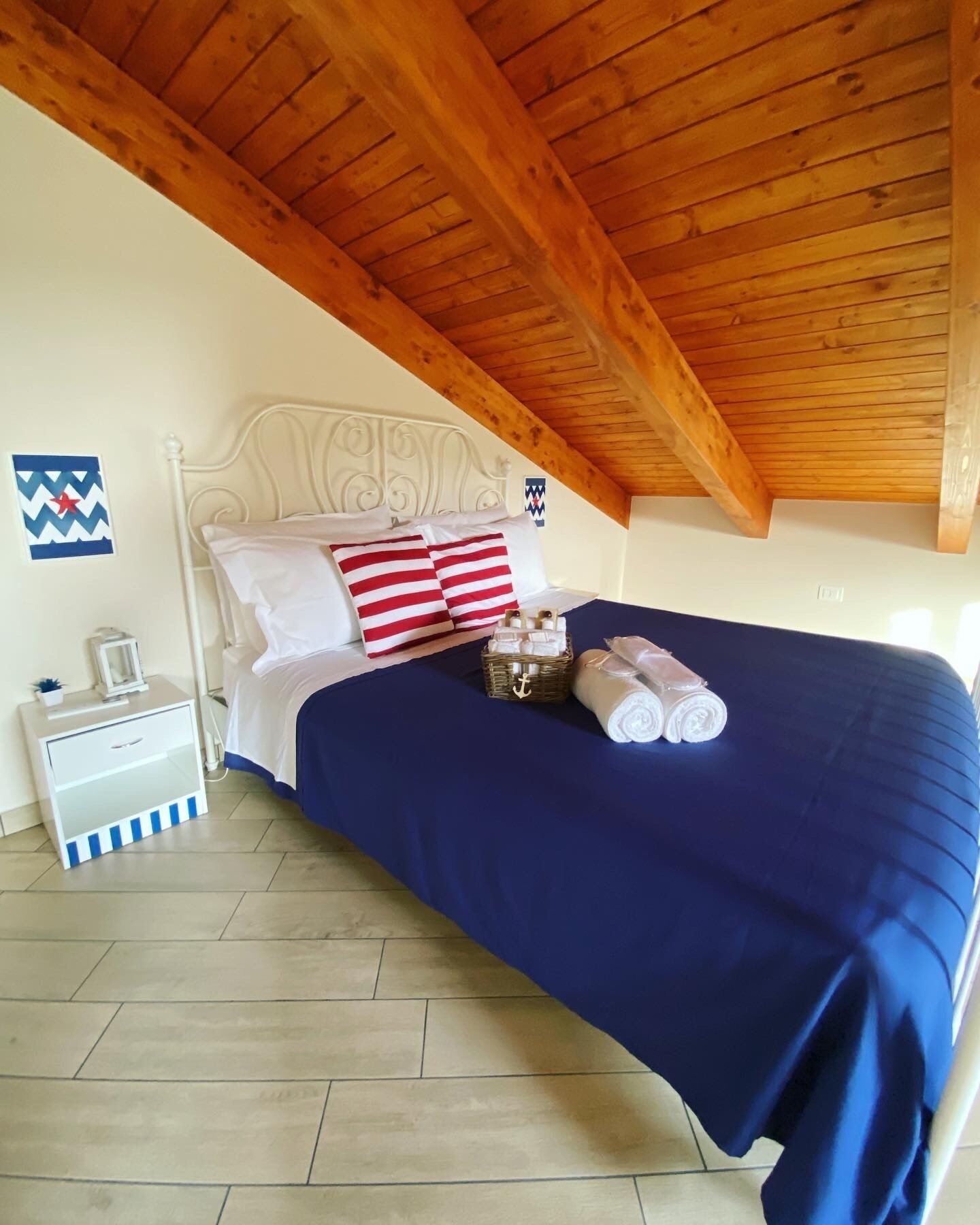 A bedroom with a wooden ceiling, a bed with a blue bedspread, white pillows, and striped pillows. There is a nightstand with a lamp and a small plant.