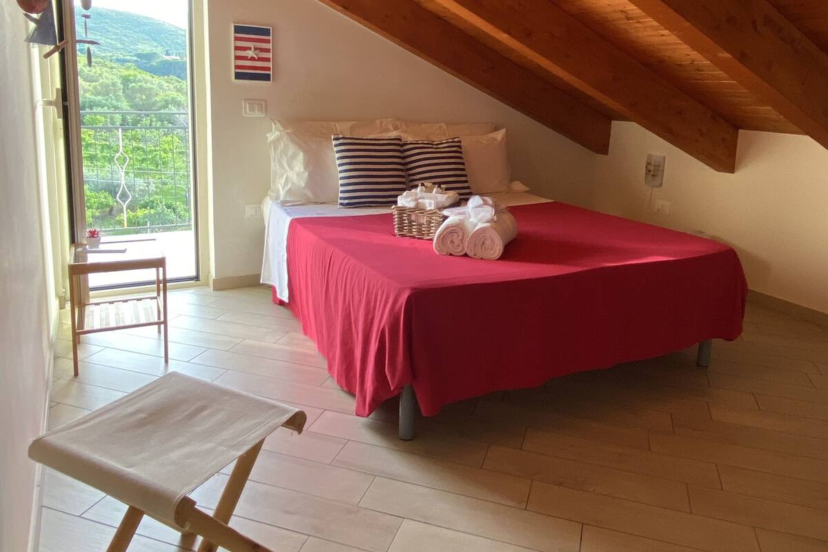 A bedroom with a sloped ceiling, a bed with a red bedspread, a small table, and a chair.