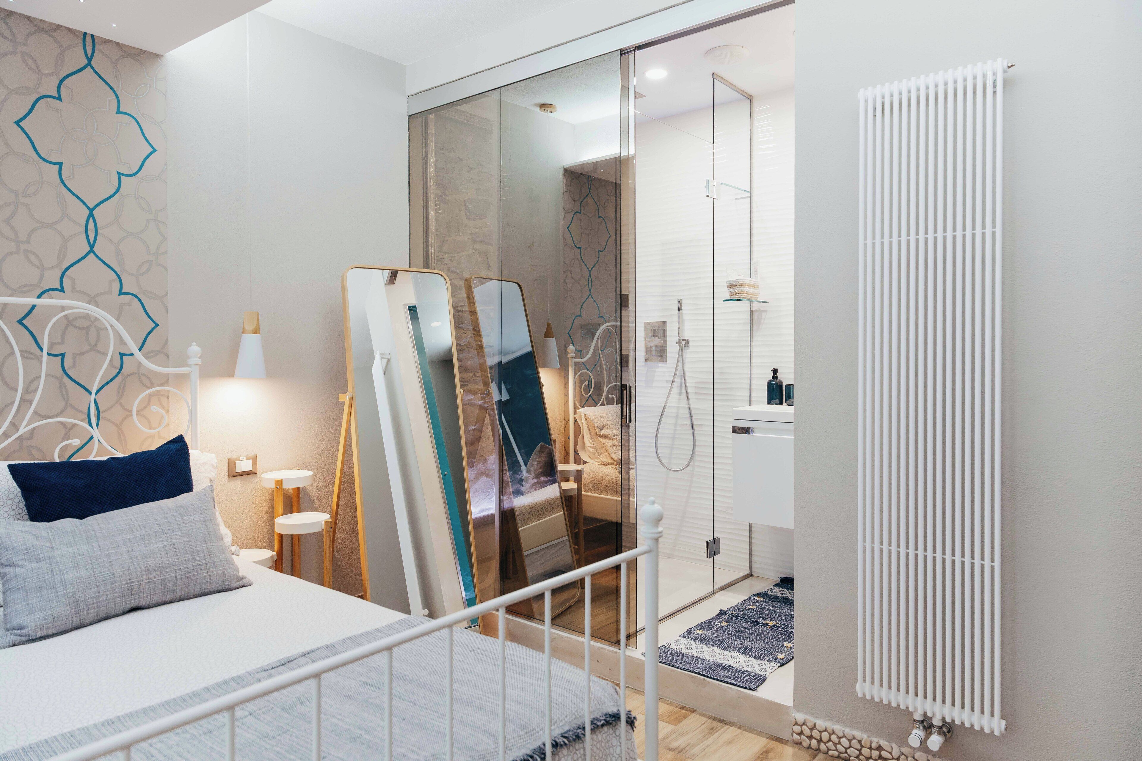 A modern hotel room with a bed, bedside tables, a mirror, a shower area, and a radiator.