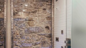 Un baño con pared de piedra, cabina de ducha de vidrio y suelo de baldosas blancas.