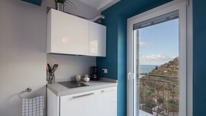 Cocina compacta con gabinetes blancos, una cafetera y utensilios sobre la encimera. Una ventana ofrece vistas de un paisaje costero.