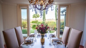 Mesa de jantar posta para seis pessoas, com um candelabro, um vaso de flores e vista para um jardim pelas janelas.