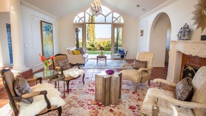 Uma sala de estar espaçosa com teto alto, grandes janelas e vista para o exterior. Há vários espaços de estar com poltronas confortáveis e uma mesa de centro.