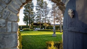 Arcada de pedra que dá acesso a um jardim bem cuidado, com uma estátua, árvores e um edifício ao fundo.