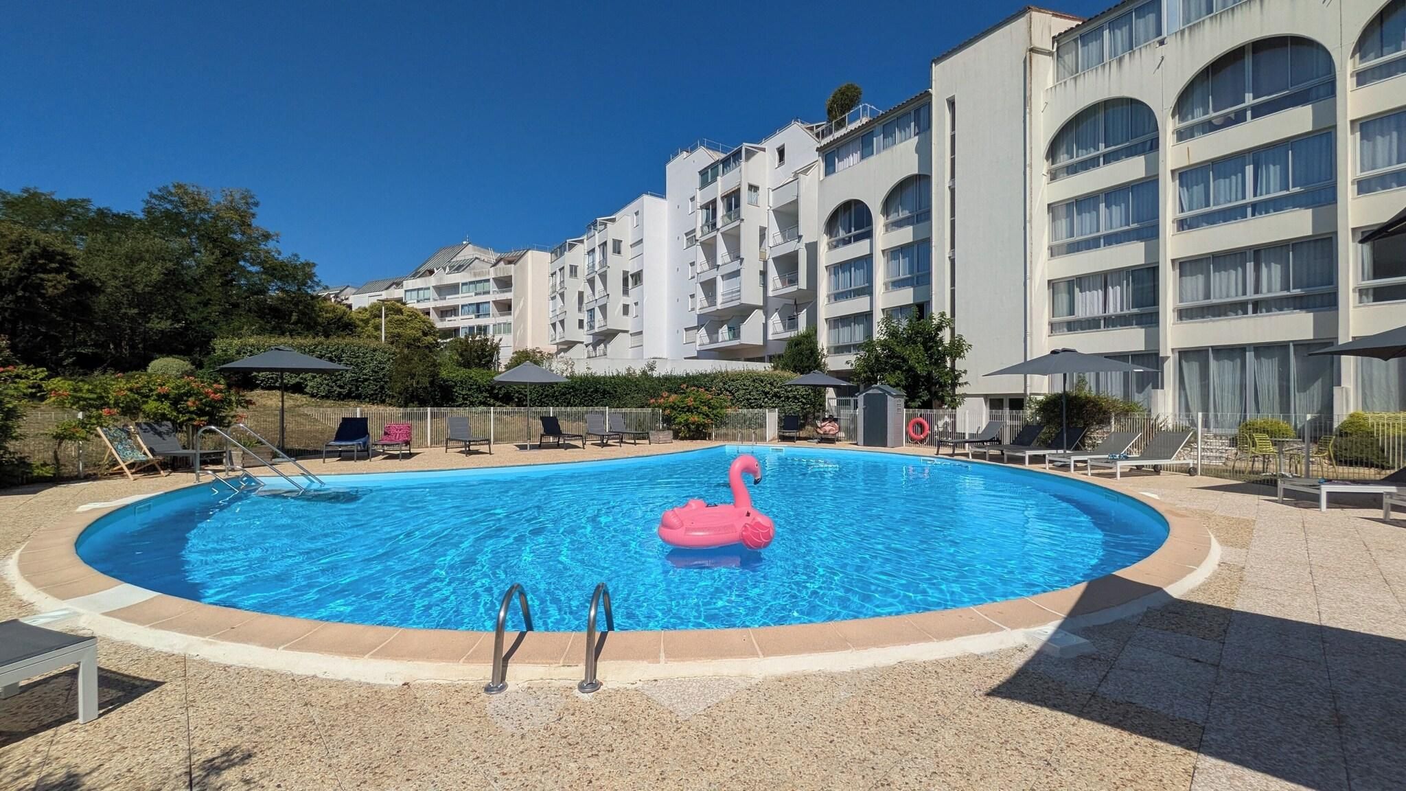 A swimming pool with a pink flamingo float, surrounded by lounge chairs and umbrellas, with a multi-story building in the background.
