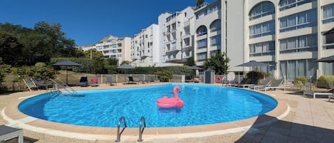 Une piscine agrémentée d’un flotteur en forme de flamant rose, entourée de chaises longues et de parasols, avec un immeuble à plusieurs étages en arrière-plan.