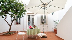 A patio with a table, chairs, and a large umbrella. There is a tree and potted plants. - GuestHost - Terrace Apartment 20 minutes from Lecce (Squinzano)