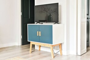 A white and blue cabinet with wooden legs, a flat-screen TV on top, and a vase with branches on the screen.