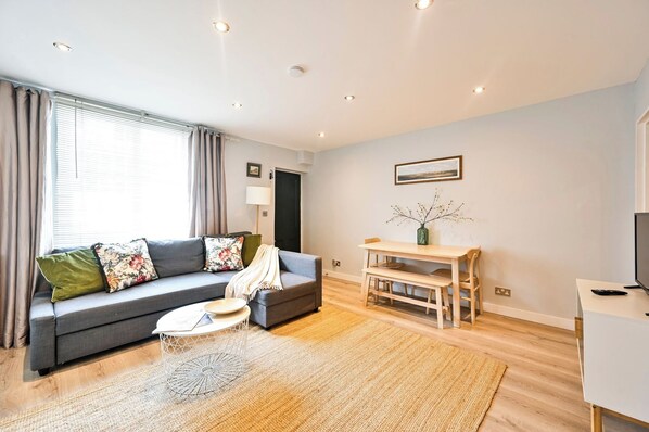 A modern living room with a grey sofa, a wooden dining table, and a TV on a white stand.