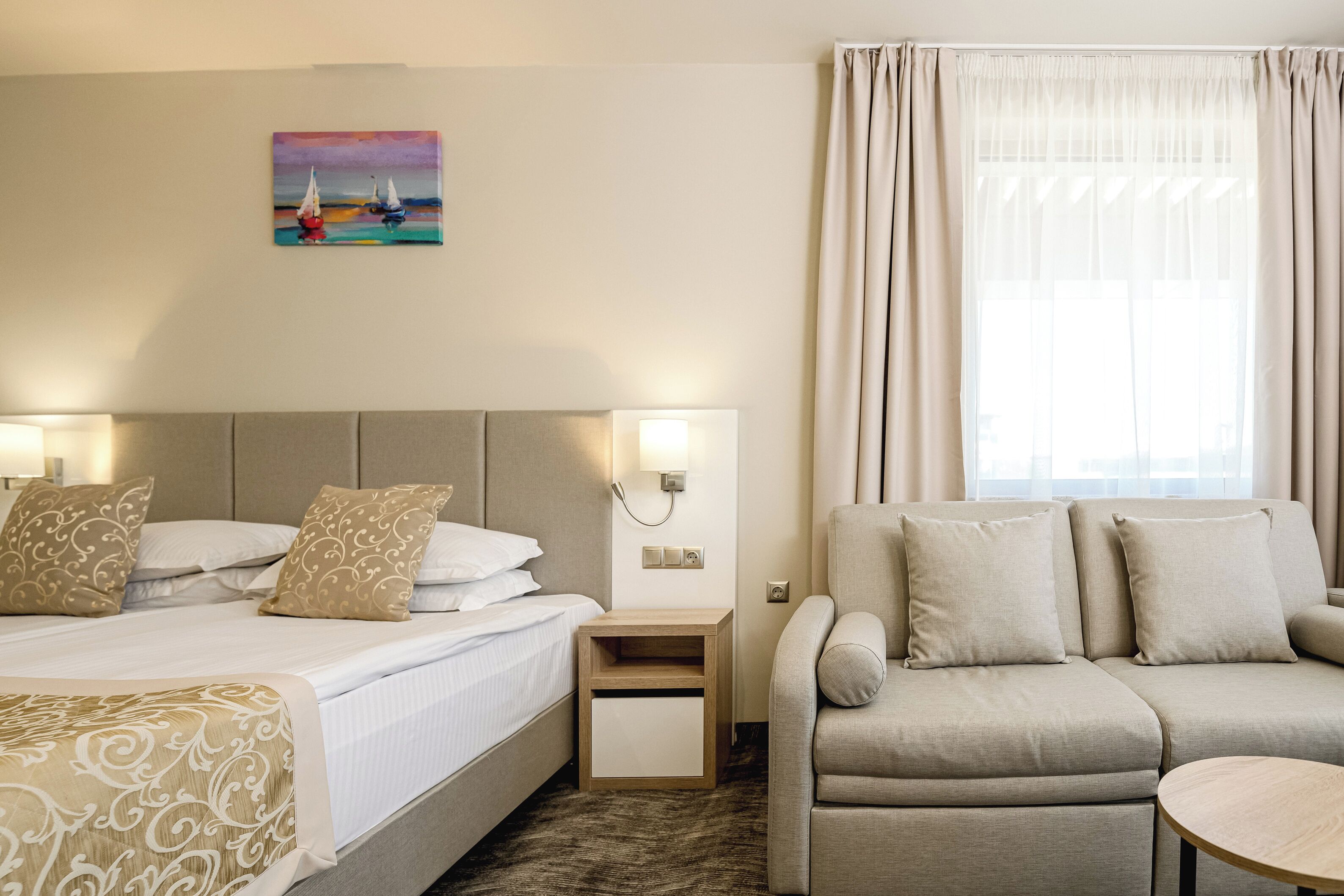 A hotel room with a bed, a sofa, a nightstand, a painting, and a window with curtains.