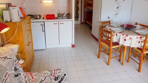 Een kleine keuken met witte kastjes, een wastafel met rode omlijsting, en een eethoek met een tafelkleed met bloemenmotief.