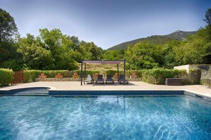 Piscina com pergolado e espreguiçadeiras, cercada por vegetação e colinas.