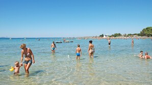 Una playa concurrida con aguas poco profundas y cristalinas, gente nadando y caminando, y una costa distante con edificios y árboles.