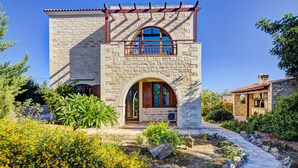 A stone building with a balcony, arched windows, and a garden with yellow flowers. - Impressive four bedroom stone house with swimming pool (Rethimno)