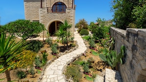 A stone building with a central arched doorway, surrounded by a garden with various plants and a stone pathway leading to the entrance. - Impressive four bedroom stone house with swimming pool (Rethimno)