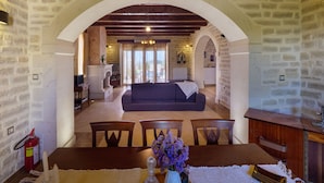 A cozy living room with a fireplace, a dining area with a table and chairs, and a kitchen in the background. - Impressive four bedroom stone house with swimming pool (Rethimno)