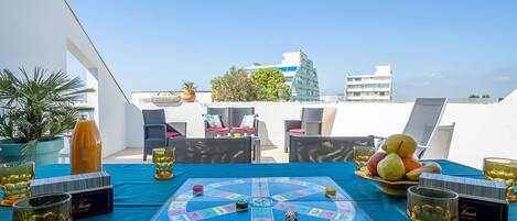Terraza en la azotea con un juego de mesa sobre un mantel azul, rodeada de mobiliario exterior y plantas en macetas.