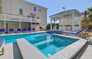 A swimming pool with lounge chairs, a covered patio, and a building with a balcony.