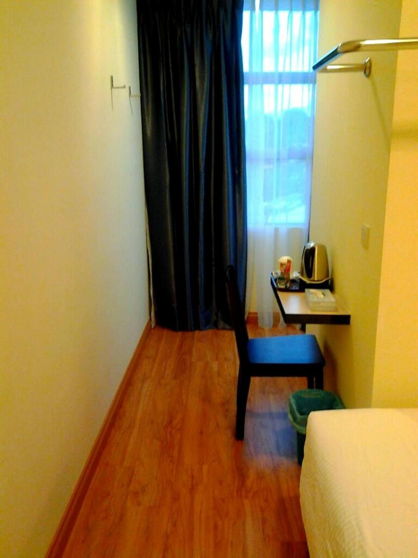 A small room with a wooden floor, a single bed, a chair, a desk with a lamp, and a window with curtains.