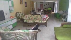 Una sala de estar con un sofá, un sillón verde y un televisor. Se observa un comedor con una mesa y sillas al fondo.