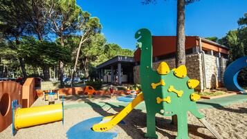 Une aire de jeux avec une structure ludique en forme de dinosaure, un toboggan jaune et une zone d’escalade bleue.
