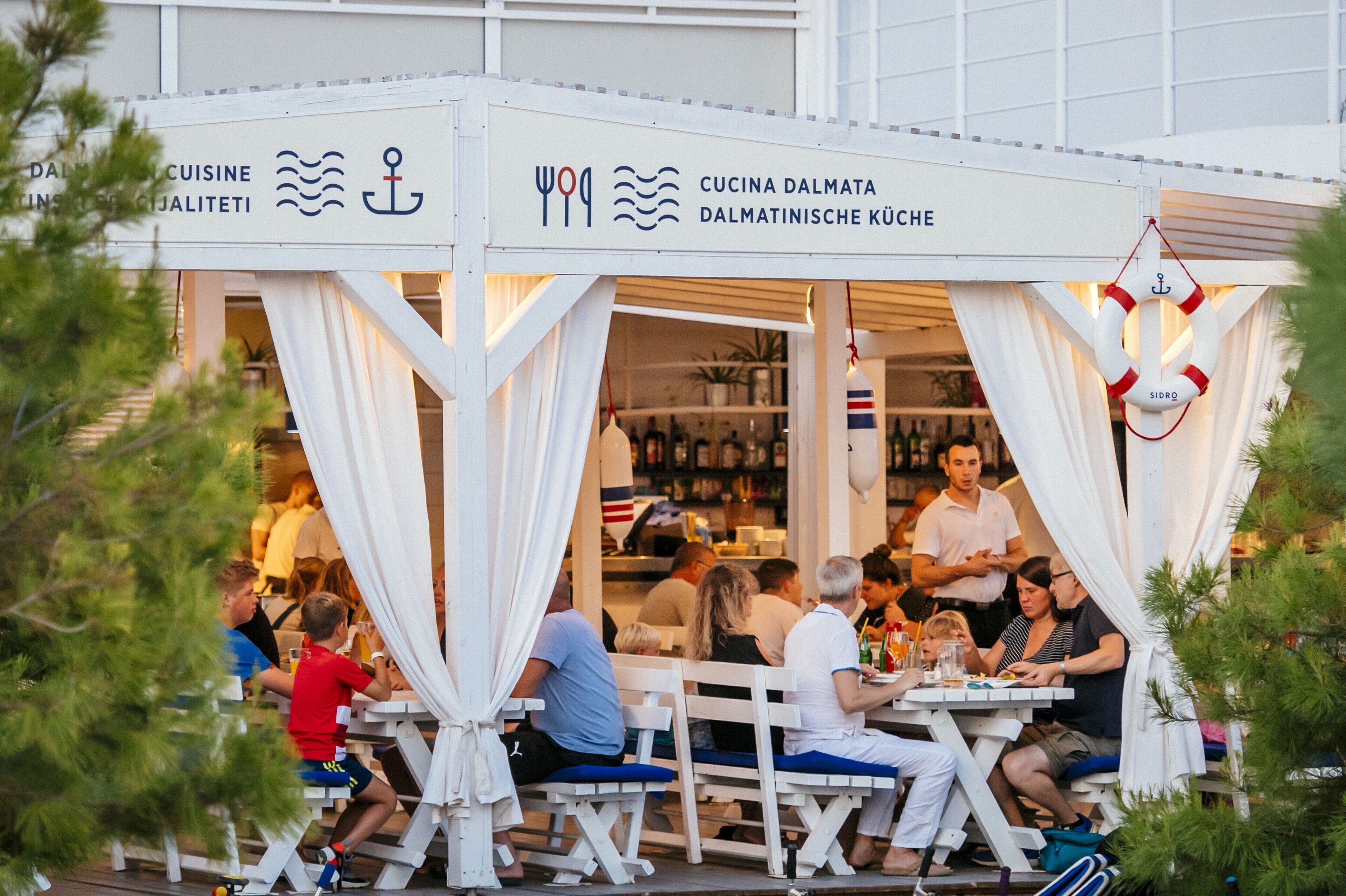 Restaurante al aire libre con toldos blancos y mesas, con temática náutica que incluye anclas y motivos de olas.