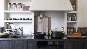Een moderne keuken met donkerhouten kasten, een inbouwoven en een groot centraal eiland. Er zijn planken met potten en keukengerei, en een hangende pannendrager boven het eiland.