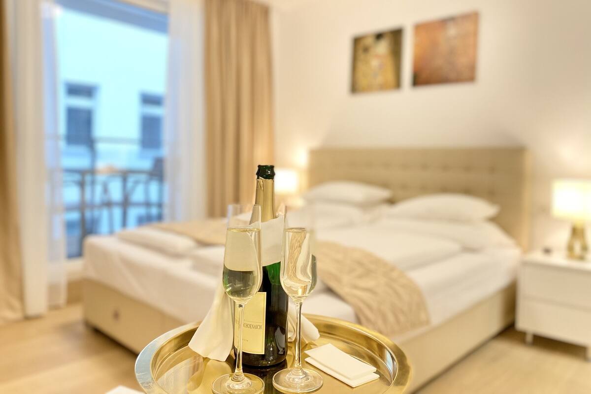 A hotel room with a bed, a bedside table with a lamp, a window with curtains, and a balcony with a railing.