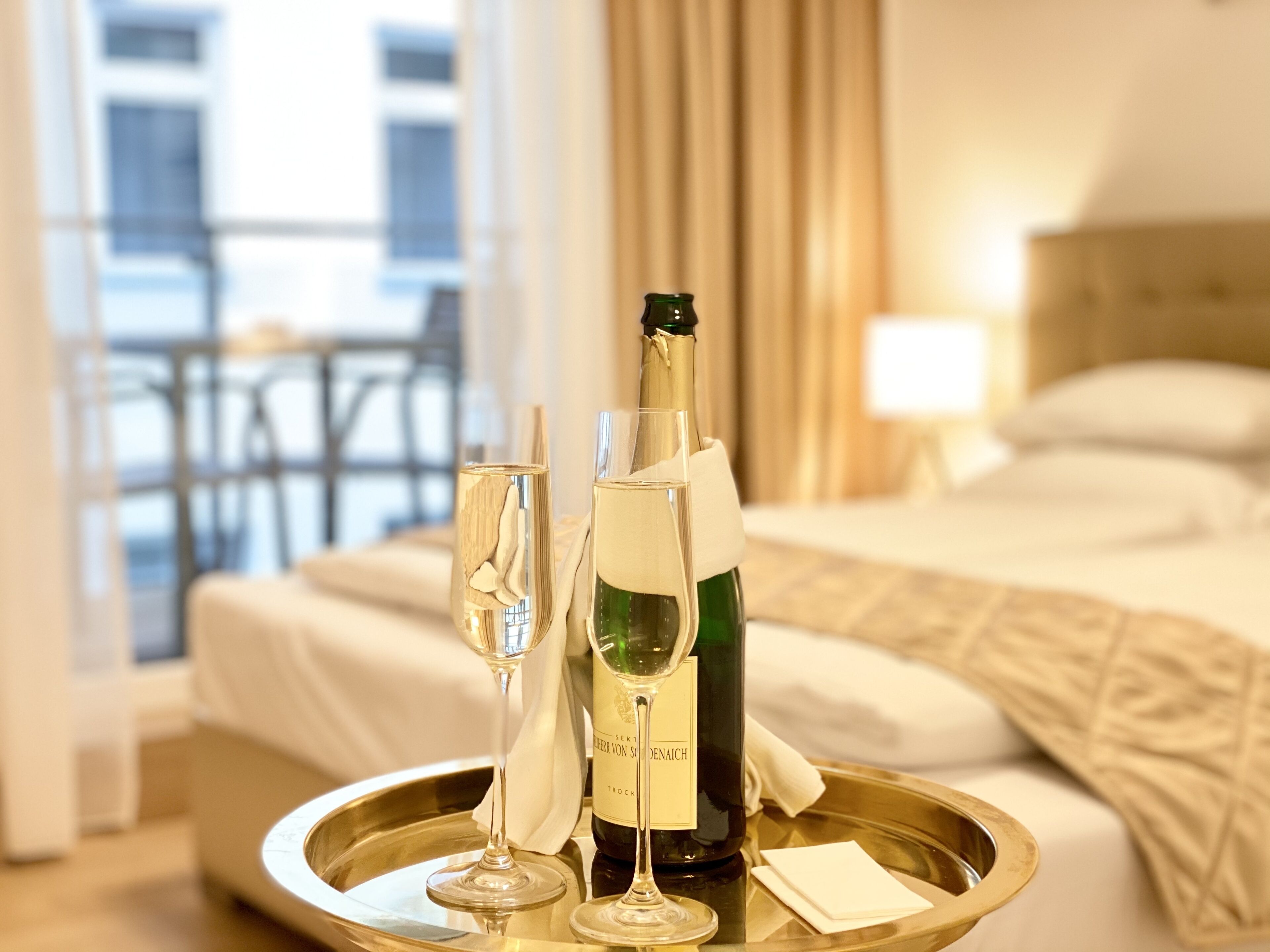 A hotel room with a bed, a bedside table, a bottle of champagne, and two champagne flutes on a tray.