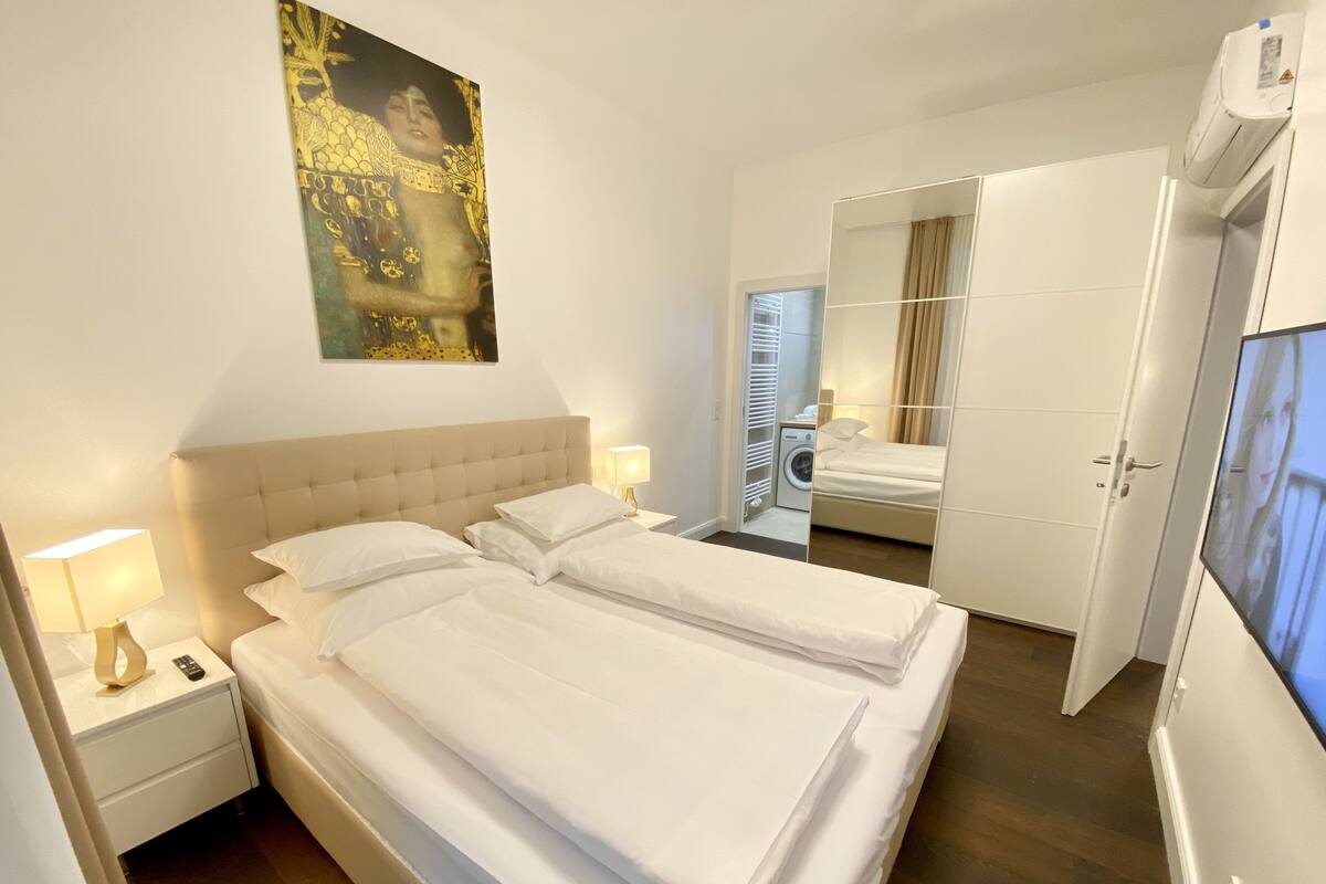A hotel room with a large bed, a bedside table with a lamp, a washing machine, and a TV.