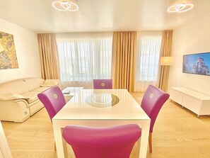 A hotel room with a glass-top dining table, purple chairs, a beige sofa, a white console, and a painting on the wall.