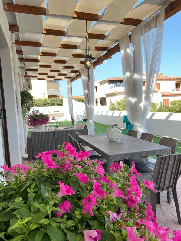 An outdoor patio with a table and chairs, surrounded by pink flowers and a pergola structure.