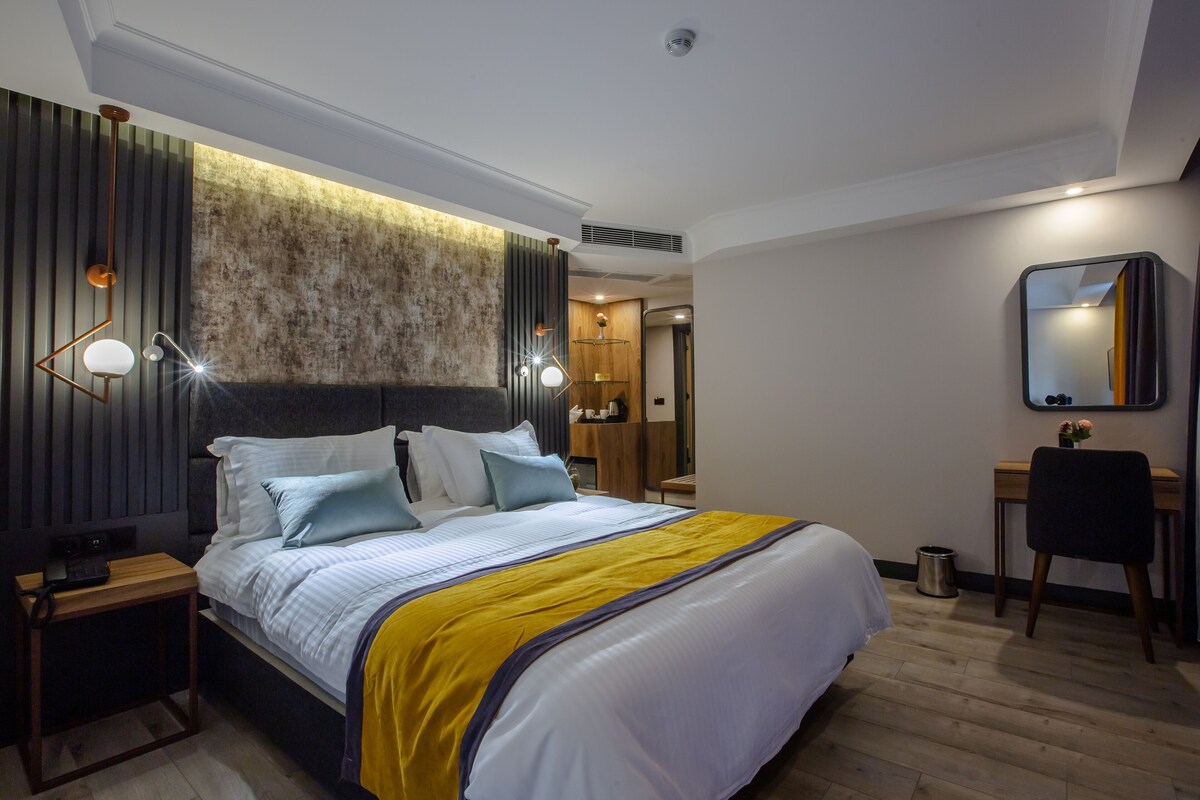 A modern hotel room with a large bed, a desk, a chair, and a mirror on the wall.